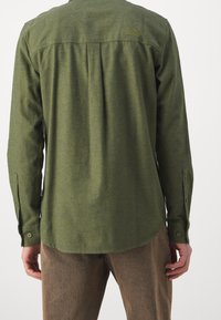 Camisa verde de manga larga con pliegue en la parte posterior. Presenta puños abotonados y un sutil detalle del logo cerca del cuello. Tejido suave y texturizado.