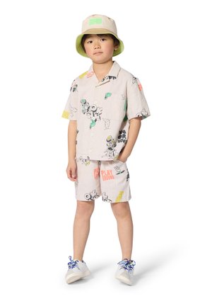 Young child wearing a beige bucket hat, matching short-sleeve button-up shirt and shorts with colorful graphic prints, and white sneakers.