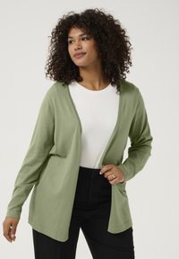 Cardigan lung cu mâneci lungi, de culoare verde deschis, cu deschidere frontală, realizat dintr-un material moale, asortat cu o cămașă albă și pantaloni negri.