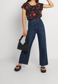 Černý květinový crop top, tmavě modré široké kalhoty s vysokým pasem, černá kabelka a sandály na platformě ukazují kontrastní textury a barvy.