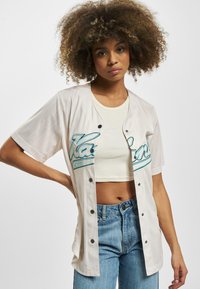 Korte ermers baseballtrøye i krem med tealfarger og vertikale striper. Har sorte knapper og en avslappet passform, brukt over en crop top.
