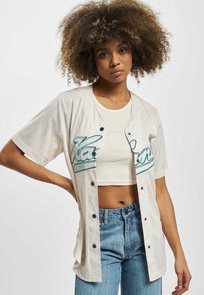 Korte ermers baseballtrøye i krem med tealfarger og vertikale striper. Har sorte knapper og en avslappet passform, brukt over en crop top.
