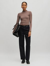 Langärmeliges, eng anliegendes braunes Oberteil mit hohem Kragen; schwarze hochsitztende Hose; schwarze glänzende Handtasche; schwarze Knöchelstiefel; gedämpfter Hintergrund.