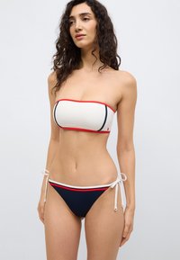 Λευκό τοπ μπικίνι bandeau με κόκκινο περίγραμμα· εσώρουχα μπικίνι σε σκούρο μπλε με λευκές και κόκκινες λεπτομέρειες, με ρυθμιζόμενα πλαϊνά κορδόνια.