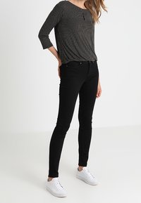 Schwarze Skinny-Jeans kombiniert mit einem schwarz-weißen gepunkteten Oberteil mit 3/4-Ärmeln und rundem Ausschnitt, ergänzt durch weiße Sneakers.