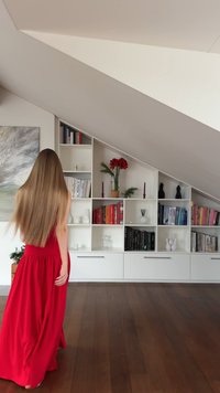 Biały regał na książki z książkami, wazą i ozdobnymi przedmiotami na tle pochyłej ściany. Drewniana podłoga. Osoba w czerwonej sukience stoi tyłem.