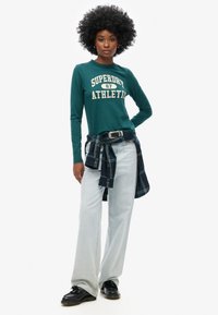 Turkusowa koszula z długim rękawem z napisem "SUPERDRY 87 ATHLETIC", jasne dżinsy oraz koszula w kratę związana w talii. Czarne buty dopełniają stylizację.