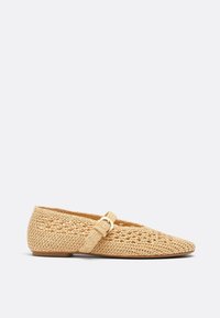 Chaussure plate tissée beige avec bout pointu et une bride unique ornée d'une petite boucle sur le dessus, sur fond uni.