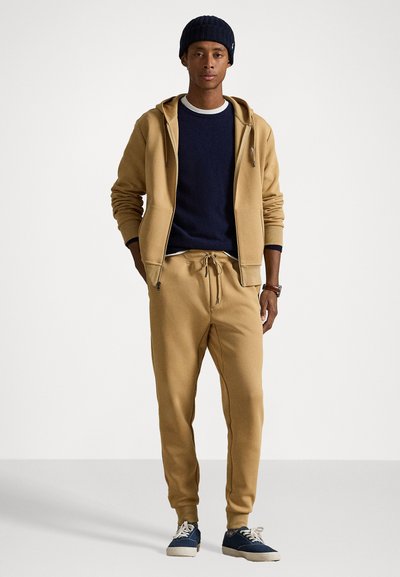 Fleece com fecho, suéter azul por baixo, calças de fato combinando e ténis azuis. Apresenta punhos canelados e cordão na cintura. Design simples.