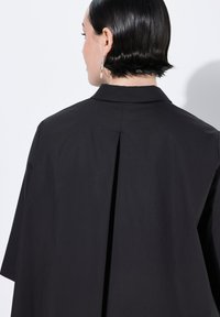 Camicia nera oversize con colletto alla francese e dettaglio a pieghe sul retro. Realizzata in tessuto liscio, presenta maniche corte e un design minimalista.