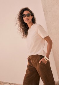 Top corto beige chiaro a maniche corte abbinato a pantaloni scamosciati marrone scuro. Gli accessori includono braccialetti in oro impilati e occhiali da sole scuri.