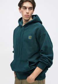Sweat à capuche bleu foncé en tissu doux, avec une poche kangourou à l'avant et un patch logo sur la poitrine. Coupe décontractée avec des poignets côtelés.