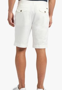 Uomo in pantaloni corti bianchi fino al ginocchio con tasche posteriori buttonate e sneakers bianche, in piedi davanti a uno sfondo neutro.