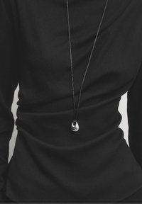Personne portant un haut noir à manches longues avec un long cordon noir en collier tenant un petit pendentif argenté brillant et irrégulier au centre.