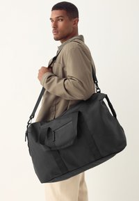 Mustast tekstureeritud kangast tehtud matkakott, millel on kaks käepidet ja reguleeritav rihm, esiküljel tõmblukuga tasku.