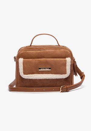 Sac en fausse suède marron avec un rabat en peluche de couleur crème, une poignée supérieure et une sangle ajustable. Présente une plaque logo dorée à l'avant.