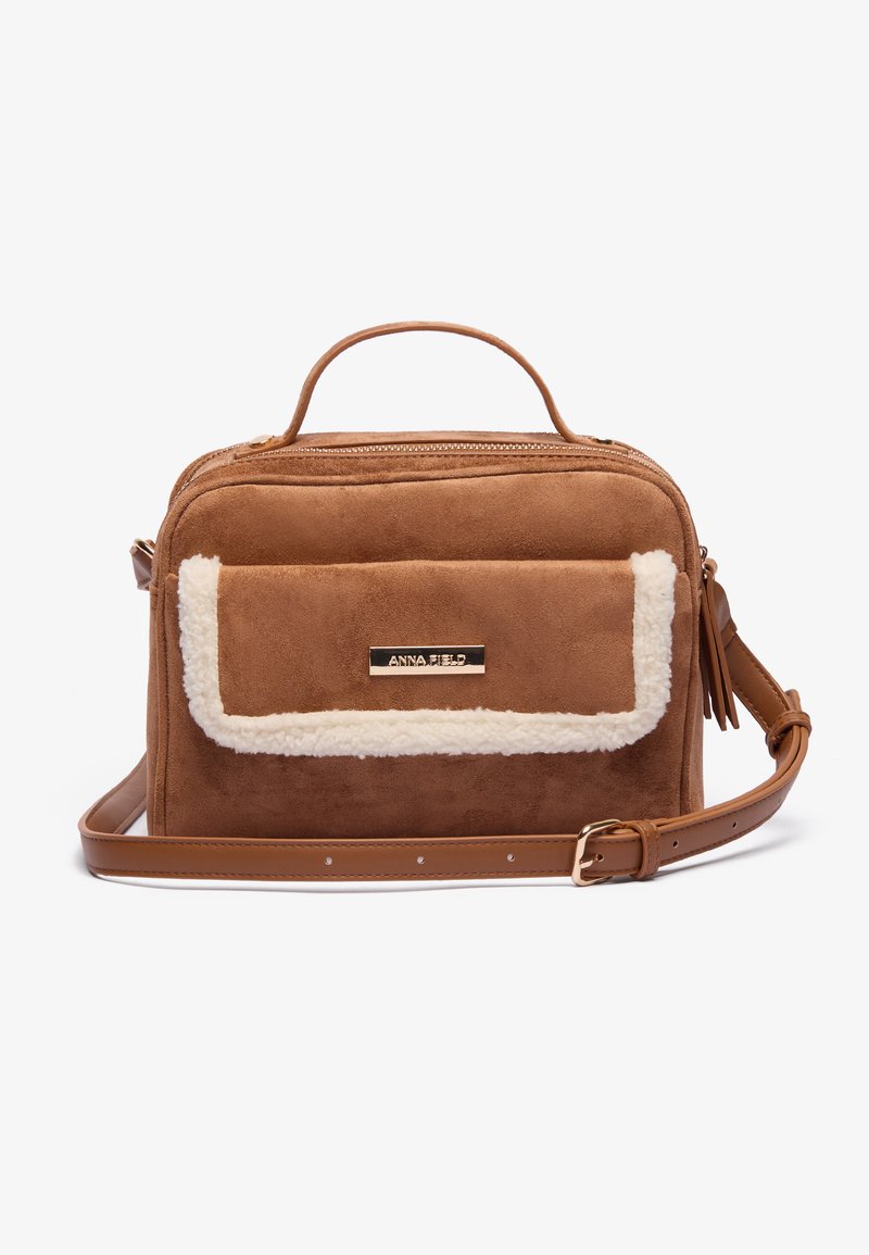 Sac en fausse suède marron avec un rabat en peluche de couleur crème, une poignée supérieure et une sangle ajustable. Présente une plaque logo dorée à l'avant.