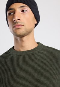 Hombre con un gorro de punto negro y un suéter de canalé verde oscuro mirando hacia un lado frente a un fondo claro y liso.