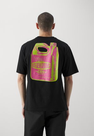 Homme portant un T-shirt noir avec un grand visuel rose et vert d’un bidon d’huile moteur et le texte « Les Benjamins » dans le dos.