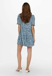 Modré květinové šaty s krátkými nafouknutými rukávy, rozšířenou sukní a nabíraným designem na zádech, doplněné bílými podpatky.