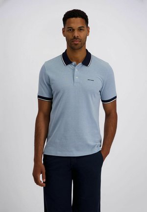 Mann steht und trägt ein hellblaues Poloshirt mit dunklem Kragen und Bündchen sowie dunkle Hosen vor einem einfarbigen Hintergrund.