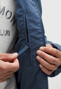 Modrá bunda s vreckom na zips na rukáve. Textile má hladkú štruktúru a zips má kontrastný modrý ťahák.