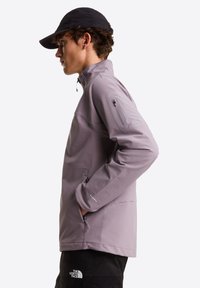 Giacca viola chiaro con mezza zip e finitura testurizzata. Presenta una tasca con zip sul braccio e tasche laterali, abbinata a un cappello nero.