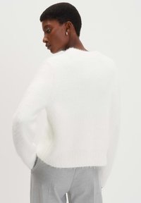 Weißer, flauschiger Pullover mit geripptem Saum und Bündchen, der eine lässige Passform bietet. Der Hintergrund ist in einer einfarbigen hellen Farbe, die das Kleidungsstück hervorhebt.