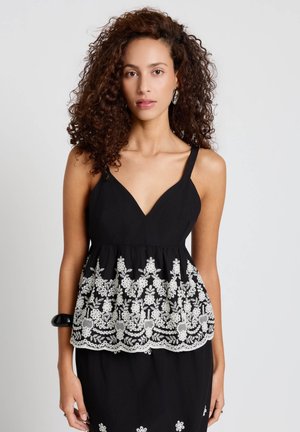 Donna con capelli ricci indossa un top nero senza maniche con ricamo floreale bianco e un braccialetto nero al polso sinistro, su uno sfondo semplice.