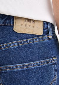 Džínsy z denimu s tmavomodrou tkaninou, viditeľným šitím a béžovou koženou nášivkou s nápisom "EDWIN" a japonskými znakmi.