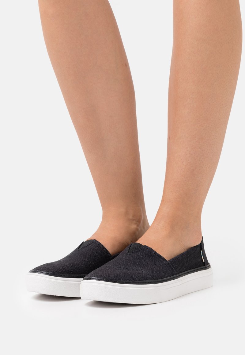 Chaussures noires à enfiler en tissu texturé, dotées d'une semelle plate blanche et de panneaux élastiques sur les côtés pour un enfilage facile. Accents de marque minimalistes visibles.