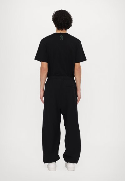 T-shirt en coton noir avec un petit motif brodé vert dans le dos, associé à un pantalon noir ample et des baskets blanches.