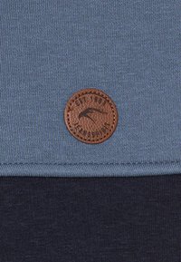 Tela azul con acabado texturizado, que presenta un parche circular de cuero marrón grabado con un pájaro y "EST. 1993." Sección inferior oscura.