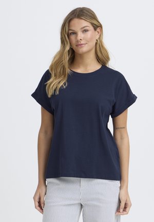 Naine, kellel on pikad blondid juuksed, kandes tumesinist lühikese varrukaga särk ja valgeid pükse peente vertikaalsete siniste triipudega, seisab tavalise tausta ees.