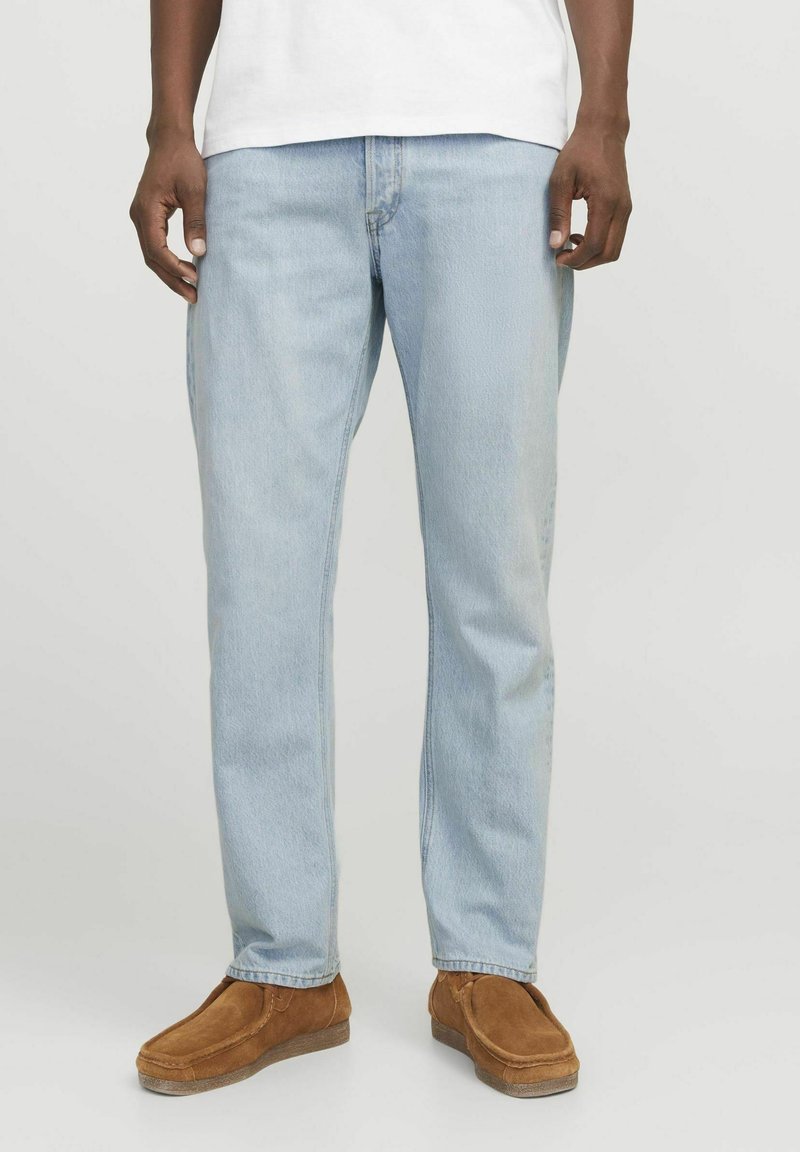 Jeans de mezclilla azul claro con corte recto, que presentan una textura desgastada y diseño de cinco bolsillos, combinados con zapatos de gamuza marrón tipo slip-on.