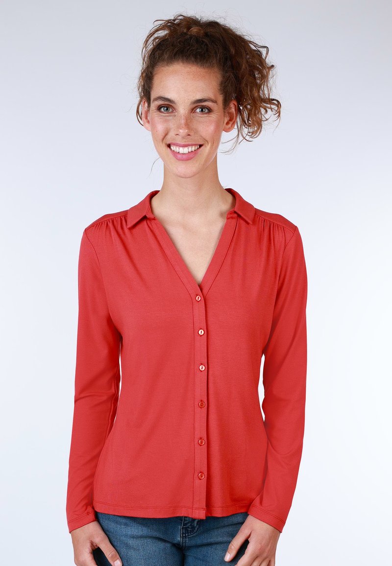 Red long-sleeve shirt with a V-neck and button-down front. Soft texture and subtle gathering at shoulders. Paired with blue jeans.