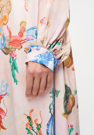 Χέρι που ακουμπάει σε ύφασμα φορέματος με θέμα τη θάλασσα, με πολύχρωμο καβούρι, ψάρι, φύκια και κύματα σε ανοιχτό ροζ φόντο.