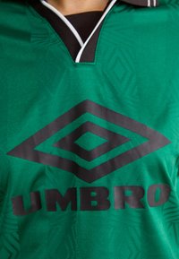 Camisa desportiva verde com um padrão texturizado. Apresenta um colarinho preto e um logótipo da Umbro em preto em destaque ao centro.