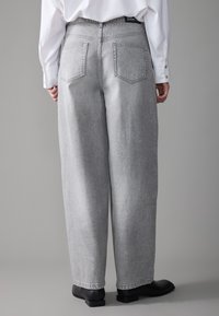 Pantalones de mezclilla grises de pierna ancha con una textura desgastada, que cuentan con dos bolsillos traseros y una etiqueta negra en la cintura, combinados con calzado negro.
