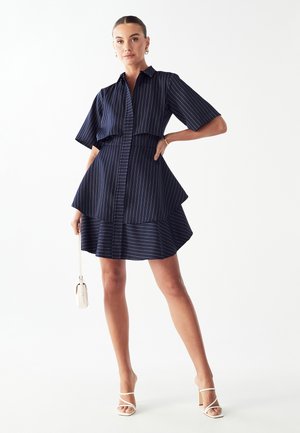 Marineblå stribet kjole med korte ærmer, knaplukning foran, lagdelt flæse i kanten og en tætsiddende talje. Sammen med hvide sandaler med stropper.