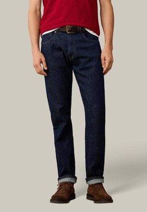 Man draagt donkerblauwe jeans met opgerolde pijpen, bruine suède schoenen, een bruine gevlochten riem en een rood shirt.