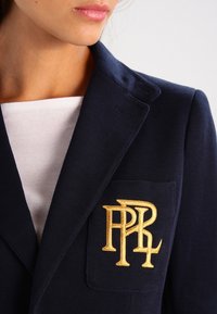 Detailní záběr na osobu ve tmavě modrém saku s vyšívanou zlatou monogramem na kapsičce a bílém žebrovaném triku pod ním.