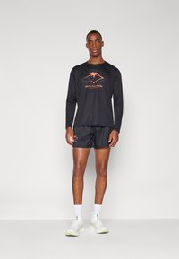 Schwarzes langärmliges Sportoberteil mit orangefarbigem Logo, kombiniert mit schwarzen Shorts. Das Modell trägt weiße Sportschuhe und Socken und steht vor einem hellen Hintergrund.