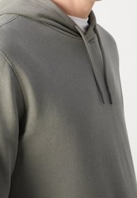 Personne portant un sweat à capuche vert olive avec des cordons sur une chemise blanche, montrée de l’épaule au cou sur un fond clair uni.