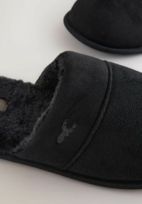 Černé pantofle vyrobené z měkké tkaniny s plyšovým vnitřkem. Jednoduchý design, plochá podrážka a vyšívané logo na boku.