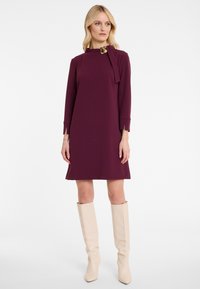 Maroon knee-length dress with long sleeves and a round neck, featuring a tie detail and golden accent. Shiny beige knee-high boots.