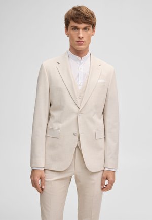 Junger Mann in einem beigefarbenen dreiteiligen Anzug und einem weißen Hemd mit Mandarin-Kragen, stehend vor einem schlichten hellgrauen Hintergrund.