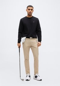 Jeune homme debout avec un club de golf, portant un pantalon beige, une chemise noire à manches longues, un gant de golf blanc glissé dans la ceinture, et des baskets blanches, sur un fond uni.