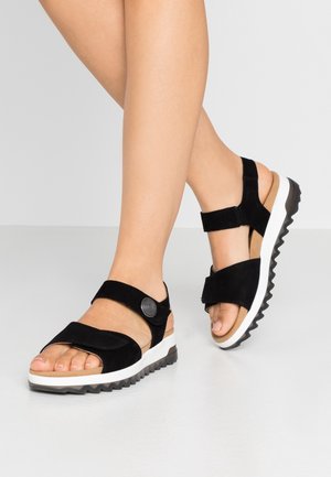 Ben iført sorte åbne sandaler med justerbare remme, polstret beige fodseng og hvide rillede såler, stående på en grå gulv.