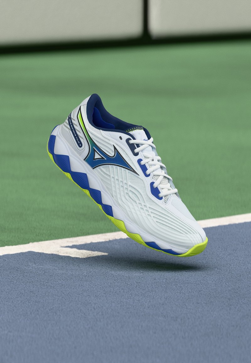 Λευκό αθλητικό παπούτσι με μπλε και νέον κίτρινες λεπτομέρειες που αιωρείται πάνω από επιφάνεια γηπέδου τένις σε αποχρώσεις μπλε και πράσινου, κοντά σε λευκή γραμμή.